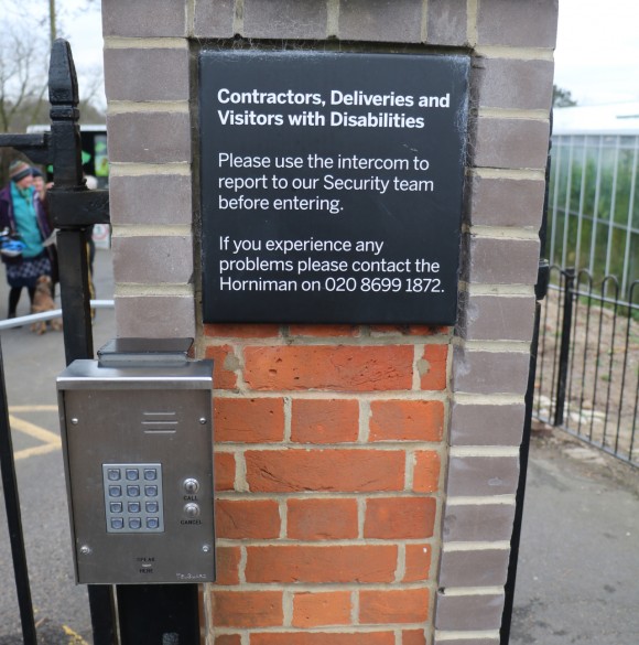 A close up of an intercom on a brick wall set in a gate (just seen). There is a sign above the intercom reading, Contractors, deliveries and visitors with disabilities. Please use the intercom to report to our security team before entering. If you experience any problems please contact the Horniman on 020 8699 1872.