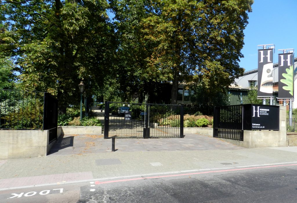 A picture taken from a road towards some black gates, which have two open sections to either side and a central closed panel. A sign to the right says Horniman Museum and Gardens entrance https-horniman-ac-uk-443.webvpn.ynu.edu.cn. There are gree trees and a path leading lighly upward in a straight line. There is an old fashion lamp just inside the gates, and some low buildings to the right